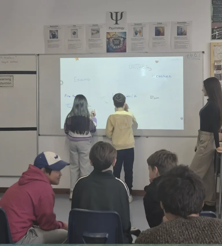 Senior psychology students at AAS Sofia lead an interactive classroom session, writing on a whiteboard while younger students observe and participate, as part of a cross-divisional collaboration on understanding and managing stress.